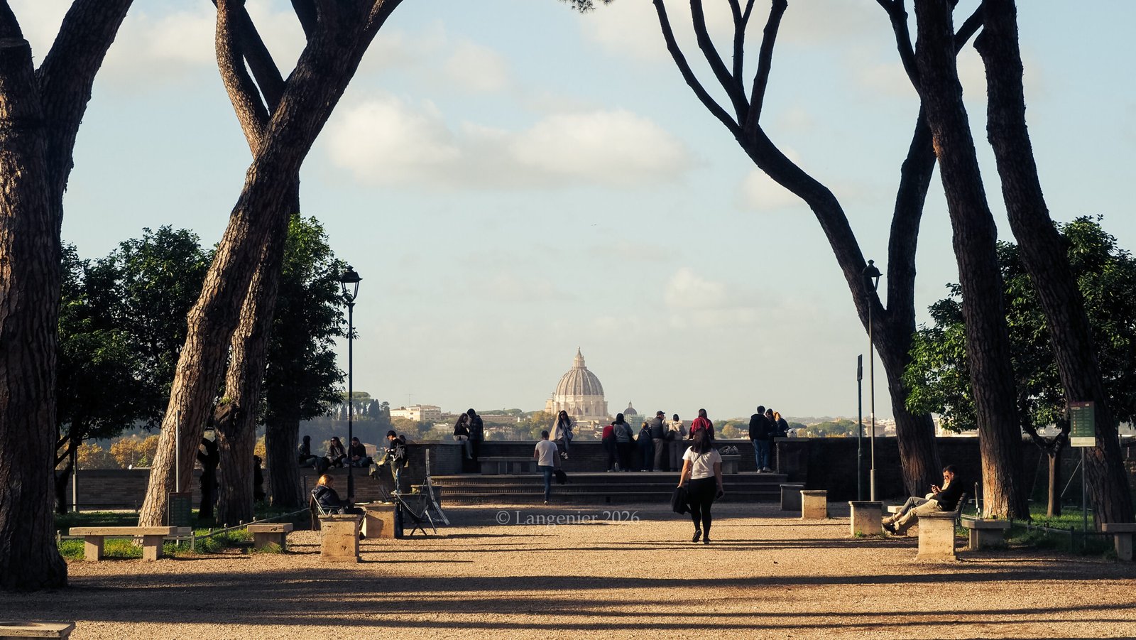 People relaxing among pine trees at the Giardino degli Aranci in Rome, with a distant view of St. Peter's Basilica dome across the city
