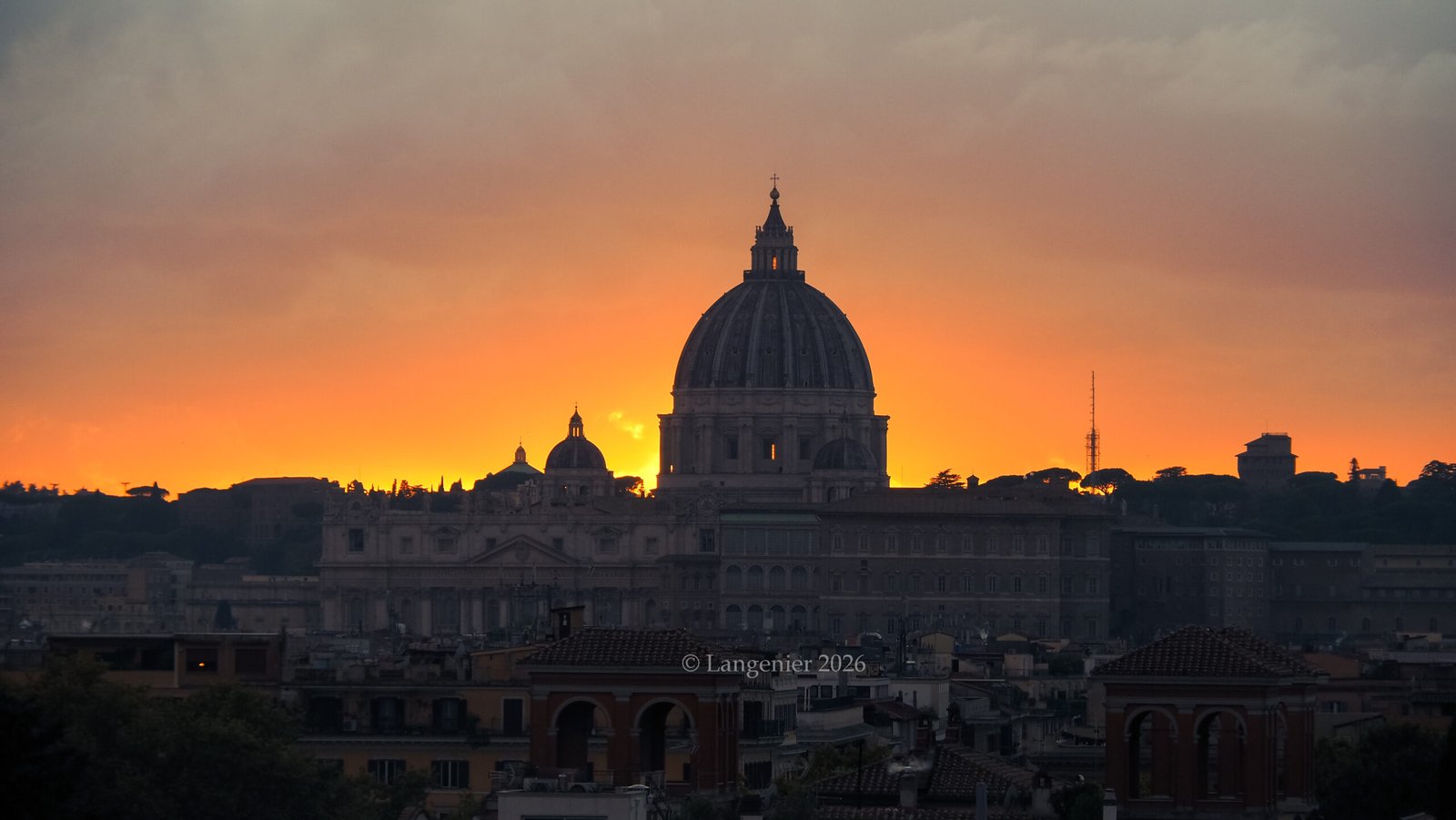 St. Peter's Basilica dome silhouetted against a fiery orange sunset, viewed from the Pincio Terrace in Villa Borghese, Rome, with the city's rooftops in the foreground.