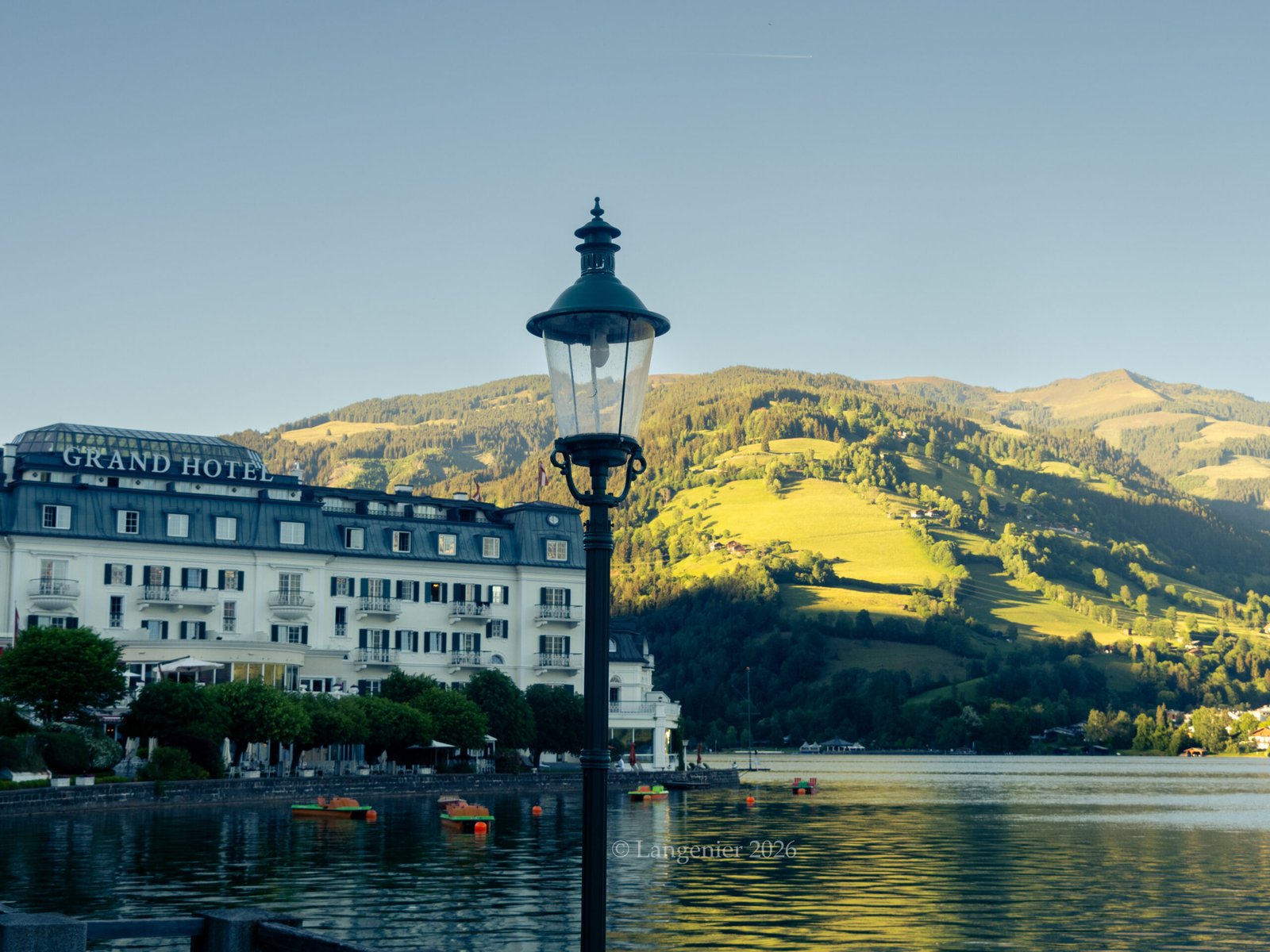 The Grand Hotel Zell am See on the shore of Lake Zell, Austria, with a wrought-iron lamppost, pedal boats on the water, and green mountains rising behind.