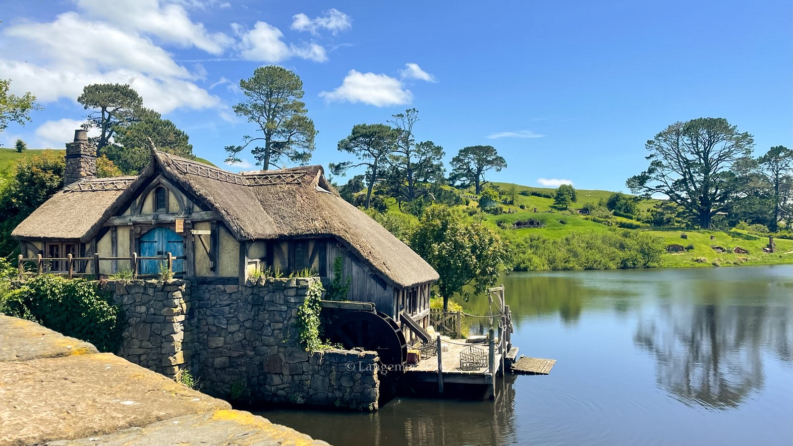 The thatched-roof watermill beside the pond at Hobbiton Movie Set near Matamata, with a stone waterwheel, wooden dock, and hobbit holes dotting the green hillside beyond.